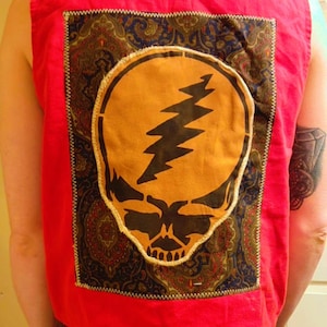 May include: Red sleeveless shirt with a large, circular patch featuring a skull and lightning bolt design. The patch is set against a paisley patterned background. The shirt is a vibrant red color.