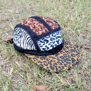 May include: A five-panel cap featuring a patchwork design of animal prints. The crown displays sections of brown leopard print, black and white zebra stripes, and a black and white spotted pattern, all separated by black fabric seams and a flat brim.