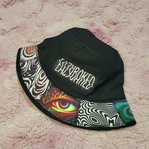 May include: A black bucket hat featuring a rectangular patch with the white text EAZYBAKED. The brim displays a colorful graphic print including a detailed human eye, swirling black and white patterns, and vibrant teal and orange abstract designs.