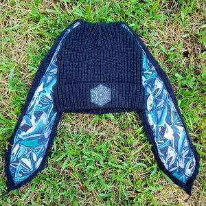 May include: A black ribbed knit beanie featuring two long, dangling side panels lined with a shiny, iridescent fabric in shades of blue, teal, and silver. A hexagonal patch with a white geometric flower of life pattern is centered on the cuff.