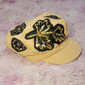 May include: A tan corduroy newsboy cap featuring black embroidered floral and leaf motifs accented with gold sequins. The hat has a structured brim and a soft, textured fabric construction with detailed stitching throughout the decorative patterns.