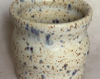 May include: A small, white ceramic cup with brown and purple speckles. The cup has a rounded shape and a slightly flared rim.