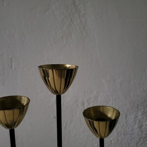 May include: A gold metal candle holder with three candle cups. The candle holder has a black metal base and three black metal stems.