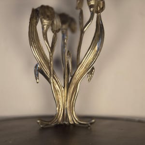 May include: A metallic silver-toned sculpture featuring multiple elongated, upright floral stems with textured leaves and bud-like tops. The structure is composed of several vertical elements that converge at a central base, creating a botanical form.