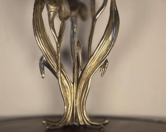 May include: A metallic silver-toned sculpture featuring multiple elongated, upright floral stems with textured leaves and bud-like tops. The structure is composed of several vertical elements that converge at a central base, creating a botanical form.