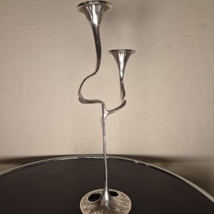 May include: A silver-toned metal candelabra featuring two candle holders at different heights. The structure consists of a single base that branches into two slender, curved stems, each topped with a flared, funnel-shaped cup for candles.