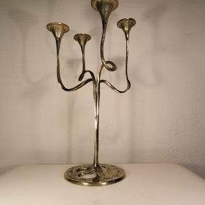 May include: A silver-colored candelabra with five candle holders. The candelabra has a round base with an ornate design. The candle holders are shaped like trumpets and are attached to curved stems.