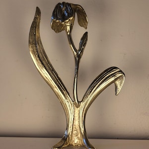 May include: A polished gold-toned metal sculpture shaped like a single flower with a drooping blossom, a slender stem, and two large, curved leaves extending from the base. The metallic surface reflects light, highlighting the textured contours.