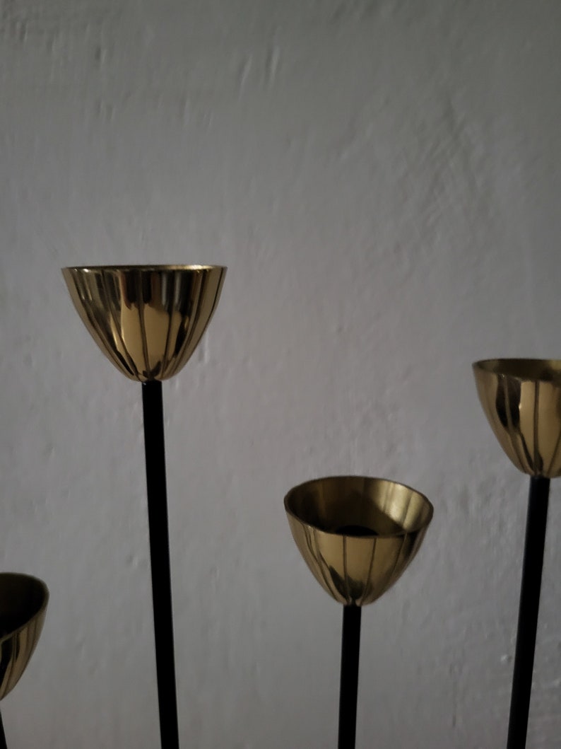 May include: A gold candle holder with five candle cups and black metal stems. The candle holder is on a white background.
