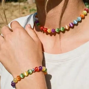 May include: A beaded necklace and a matching beaded bracelet featuring round, multi-coloured cat's eye beads in shades of red, orange, yellow, green, blue, and purple. The jewellery set includes small silver-toned spacer beads between the stones.