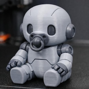 May include: A grey 3D-printed robot baby toy with a dummy. The robot has a rounded head, large eye sockets, and articulated limbs. The toy is sitting down, with its arms and legs bent.