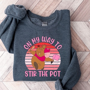 May include: A dark gray sweatshirt with a cartoon pig wearing a pink hat and holding a pink spoon. The text "On my way to stir the pot" is printed on the sweatshirt.