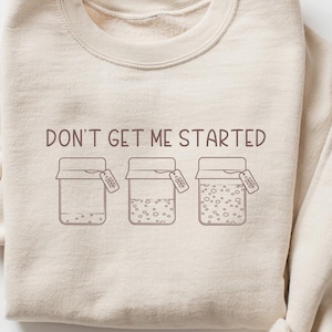 May include: A beige sweatshirt with the text "DON'T GET ME STARTED" and a graphic of three jars with lids.