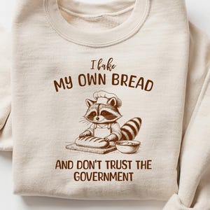 May include: A beige sweatshirt with the text "I bake my own bread and don't trust the government" and a cartoon raccoon wearing a chef's hat and baking bread.