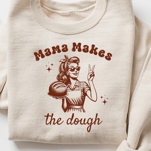 May include: A beige sweatshirt with a brown graphic of a woman holding a loaf of bread and the text "Mama Makes the Dough".