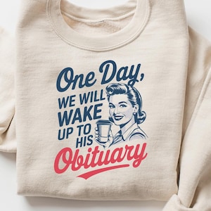 Puede incluir: Sudadera color crema con el texto "One Day, We Will Wake Up to His Obituary" en azul y rojo. Presenta una ilustraci&oacute;n de estilo vintage de una mujer sosteniendo una taza.