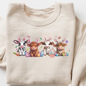 May include: A beige sweatshirt with a graphic of four cows wearing bunny ears and holding Easter eggs. The cows are black and white, brown, and white, and brown and white. The graphic is surrounded by flowers and butterflies.