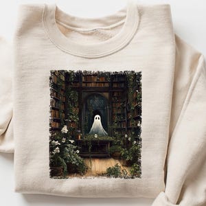 May include: Cream-colored sweatshirt featuring a graphic of a ghost in a library setting. The artwork depicts a spectral figure in a doorway surrounded by bookshelves and greenery. The sweatshirt has a crew neck and long sleeves.