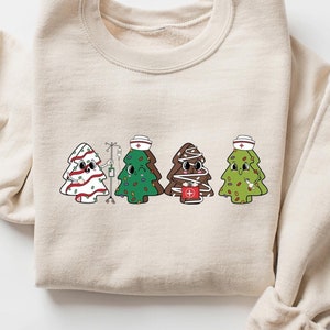 May include: A beige sweatshirt with a graphic of four Christmas trees dressed as nurses. The trees are wearing red and white striped hats, green hats, and brown hats. One tree is holding a red cross.