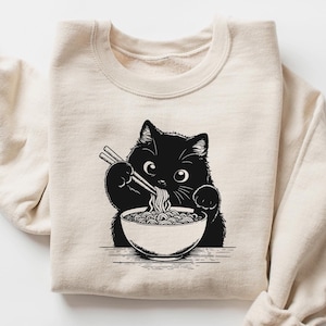 May include: A beige sweatshirt with a black cat eating ramen noodles from a bowl with chopsticks. The cat is looking at the viewer.
