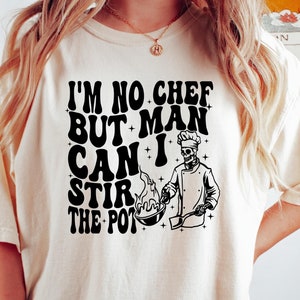 May include: A white t-shirt with a black graphic of a skeleton chef cooking in a wok. The text on the shirt reads "I'm no chef but man can I stir the pot."