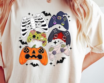 Op de afbeelding: Een crèmekleurig T-shirt met een afbeelding van vijf verschillende Halloween-videogamecontrollers. De controllers zijn versierd met skeletten, mummies, pompoenen en andere Halloween-afbeeldingen.