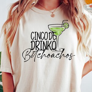 以下が含まれることがあります： マルガリータグラスと「Cinco de Drinko Bitchoachos」というテキストの黒いグラフィックが描かれた白いTシャツです。