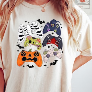 May include: A cream colored t-shirt with a graphic of five different Halloween themed video game controllers. The controllers are decorated with skeletons, mummies, pumpkins, and other Halloween imagery.