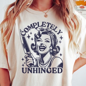 May include: A cream colored t-shirt with a graphic of a woman holding a knife and the text "COMPLETELY UNHINGED".