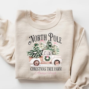 May include: A beige sweatshirt with a Christmas tree farm design. The design features a white truck with a Christmas tree on the back, and the text "North Pole Christmas Tree Farm Always Fresh".