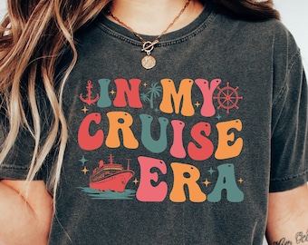 Puede incluir: Una camiseta gris oscuro con un gráfico de estilo retro que dice "In My Cruise Era". El gráfico presenta un crucero, palmeras, un ancla y un timón.