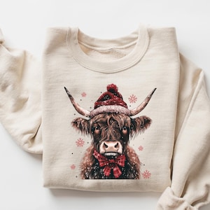 May include: A beige sweatshirt with a brown and white highland cow wearing a red Santa hat and a red bow tie. The cow is surrounded by small pink snowflakes.