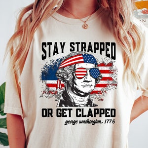 May include: A white t-shirt with a graphic of George Washington wearing sunglasses and a bandana with the American flag. The text "Stay Strapped or Get Clapped" is printed on the shirt, along with the text "george washington. 1776".