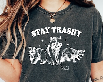 May include: A dark grey t-shirt with a white graphic of raccoons wearing sunglasses and the text "Stay Trashy".