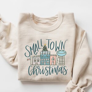 May include: A beige sweatshirt with a graphic design of a small town with a Christmas tree and the text "Small Town Christmas".