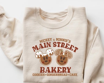 May include: A beige sweatshirt with a brown graphic design that says "Mickey & Minnie's Main Street Bakery" with images of Mickey and Minnie Mouse as gingerbread cookies. The text below the graphic says "Cookies, Gingerbread, Cake."