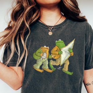 May include: A dark gray t-shirt with a graphic print of two frogs. One frog is holding a ball of yarn and the other is holding a kite. The text "More B&eacute;b&eacute;s" is visible on the right arm.