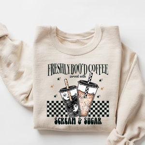 以下が含まれることがあります： ゴーストが描かれたコーヒーカップ2つと「Freshly Boo'd Coffee served with Scream & Sugar」というテキストがデザインされたベージュのトレーナーです。