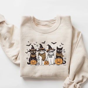 May include: A beige sweatshirt with a graphic print of three cats wearing witch hats and holding pumpkins. The cats are black, white, and gray. The graphic is in a cartoon style.
