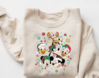 Puede incluir: Una sudadera beige con un diseño navideño con Mickey Mouse, Minnie Mouse, Donald Duck y Goofy con gorros de Papá Noel y rodeados de luces navideñas.