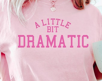 Könnte beinhalten: Ein pinkfarbenes T-Shirt mit dem Text "A Little Bit Dramatic" in pinkfarbener Schrift. Die Person trägt blaue Shorts und hält eine weiße Tasse mit einem gelben Smiley-Gesicht.