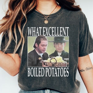 May include: A dark gray t-shirt with a white graphic print of two men in colonial attire. The text "WHAT EXCELLENT BOILED POTATOES" is printed below the image.