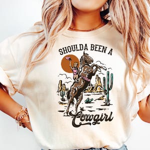 以下が含まれることがあります： クリーム色のTシャツを着た女性。Tシャツには「Shoulda Been a Cowgirl」という文字と、砂漠の風景の中で馬に乗っている女性のグラフィックが描かれている。彼女は青いジーンズも履いている。