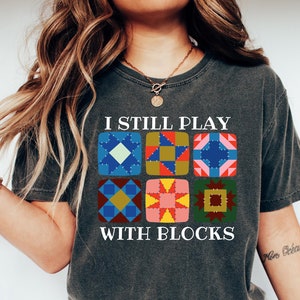 May include: A dark gray t-shirt with the text "I still play with blocks" and a graphic of nine colorful patchwork quilt blocks.