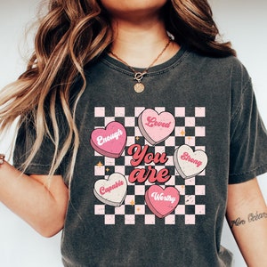May include: A gray t-shirt with a checkered pattern and pink hearts that say "You are" with words like "Loved", "Strong", "Enough", "Capable", and "Worthy" inside the hearts.