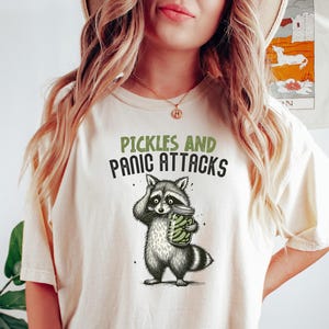 May include: Cream-colored t-shirt with the text "Pickles and Panic Attacks" above a cartoon raccoon holding a jar of pickles. The raccoon is black, white, and gray. The shirt is worn by a person.