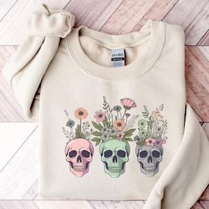 May include: A beige sweatshirt with a graphic of three colorful skulls with flowers growing out of the top of their heads.