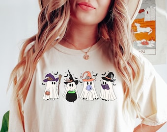 Può includere: Una maglietta bianca con una grafica di quattro fantasmi che indossano cappelli da strega. I fantasmi tengono una zucca, un calderone, una luna e un libro. La maglietta è di colore crema chiaro e ha una vestibilità rilassata.