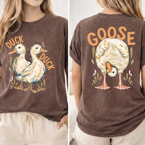 Puede incluir: Camiseta marr&oacute;n con un gr&aacute;fico de pato en la parte delantera y un gr&aacute;fico de ganso en la parte trasera. La parte delantera de la camiseta presenta dos patos con la palabra "DUCK" arriba y abajo. La parte trasera de la camiseta tiene un ganso con la palabra "GOOSE" arriba.