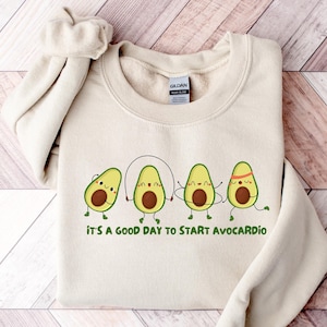 Könnte beinhalten: Ein beiger Pullover mit einer Grafik von vier Cartoon-Avocados, die Seilspringen. Der Text "It's a good day to start avo-cardio" ist unter den Avocados gedruckt.